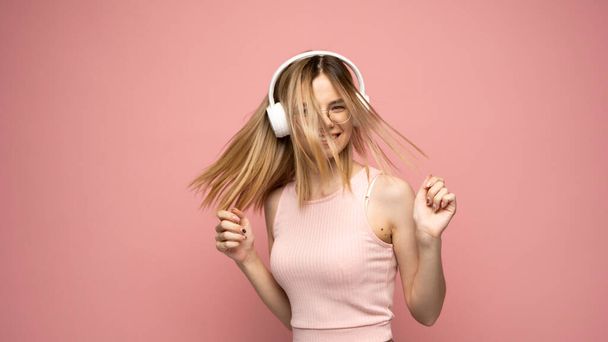 Beautiful attractive young blond woman wearing pink t-shirt and glasses in white headphones listening music and dancing on pink background in studio. Relaxing and enjoying. Lifestyle. - Фото, зображення