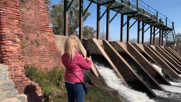 Kobieta filmująca za pomocą smartfona odpływ wody z bram zapory w San Antonio de Areco. Koncepcja podróży - Materiał filmowy, wideo