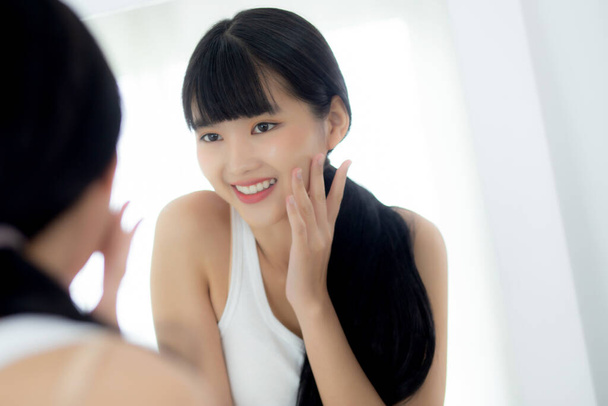 Krásná tvář mladé asijské ženy se šťastným úsměvem a dotek tvář hledá zrcadlo s okouzlující, make-up krásy obličeje dívka s péčí o pleť a kosmetické ošetření, péče o pleť a zdravý koncept. - Fotografie, Obrázek