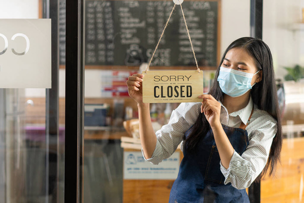 Πορτρέτο ενός όμορφου Ασιάτη μπαρίστα με ποδιά. Κρατώντας μια πινακίδα με τη λέξη κλειστή στην γυάλινη πόρτα που εργάζονται σε μια καφετέρια. - Φωτογραφία, εικόνα