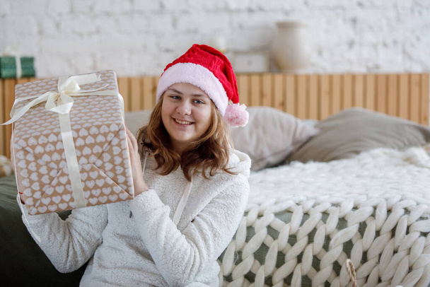 Szőke tinédzser lány karácsonyi ajándékkal egy dobozban ül a padlón a kanapé mellett. Vörös Mikulás kalap. Szelektív fókusz. - Fotó, kép