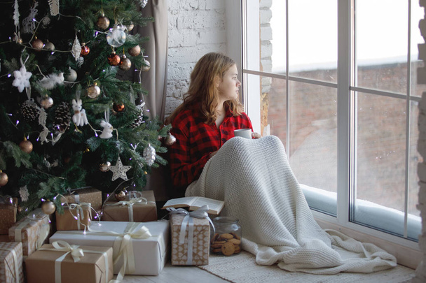 Blonde teenager girl sits near a Christmas tree by the window. Red plaid shirt, long hair, dreamy look. Merry Christmas. Selective focus. - Photo, Image