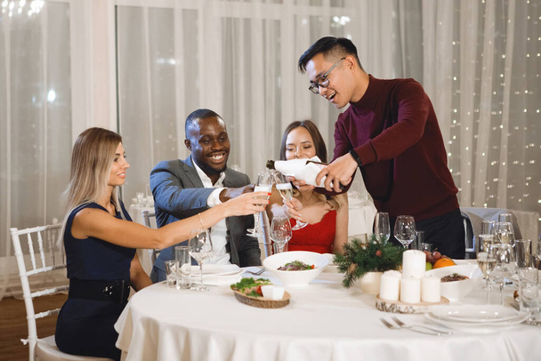 Společnost nadnárodních přátel u slavnostního stolu s sklenicemi šampaňského, usmívající se a hovořící v restauraci. Selektivní zaměření, rozmazaná světla. - Fotografie, Obrázek