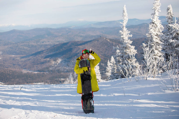 Mulher snowboarder em roupas de esqui posa com uma placa contra um fundo de montanhas azuis. Casaco amarelo com capuz, chapéu de malha, óculos de esqui. Estilo de vida saudável. Conceito desportivo. Foco seletivo. - Foto, Imagem