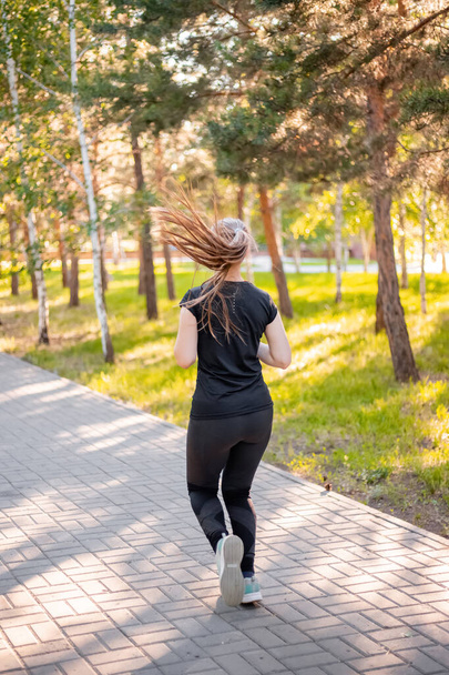 Back view a girl in a black sports uniform is jogging in the evening park. Self-sports concept.  - Foto, afbeelding