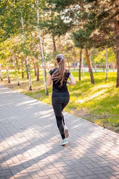 Back view a girl in a black sports uniform is jogging in the evening park. Self-sports concept.  - Fotó, kép