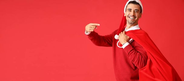 Junger Mann mit Weihnachtsmütze hält Tasche voller Geschenke auf rotem Hintergrund mit Platz für Text - Foto, Bild