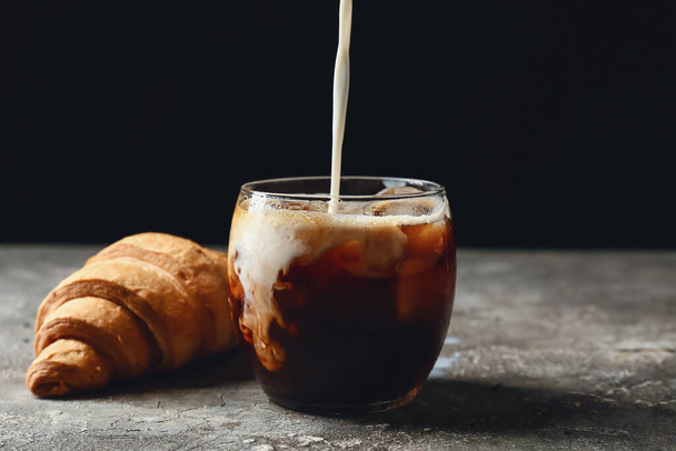 Наливание молока в стакан с вкусным кофе со льдом и круассаном на стол - Фото, изображение