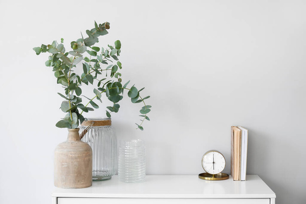 Vases with eucalyptus branches, alarm clock and books on shelf near light wall - Photo, image