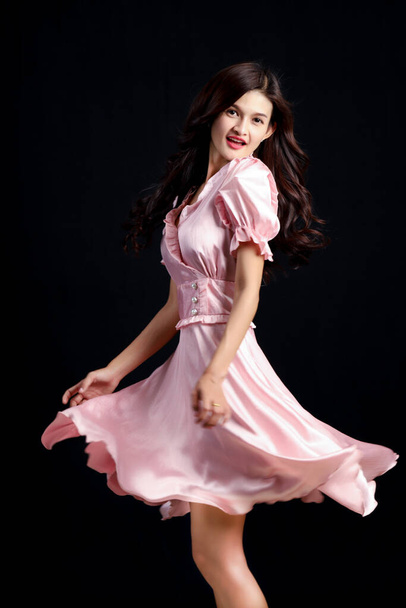 Портрет улыбающейся красивой женщины, разворачивающейся и смотрящей в камеру на черном фоне, молодой азиатки позирует в трепещущем розовом платье в студии. - Фото, изображение