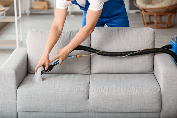 Male worker removing dirt from grey sofa with vacuum cleaner in room - 写真・画像