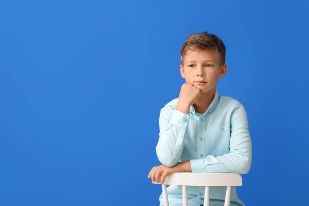 Bedachtzame kleine jongen zittend in stoel op blauwe achtergrond - Foto, afbeelding