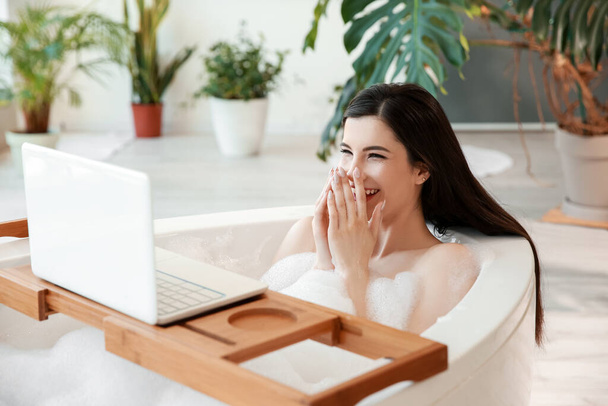 Mujer bastante joven viendo vídeo en el ordenador portátil mientras toma un baño en casa - Foto, imagen