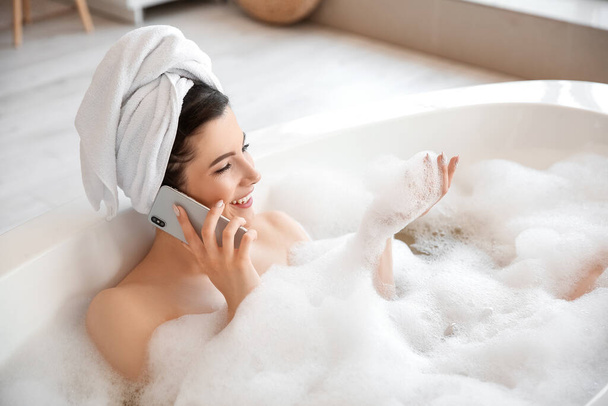 Ευτυχισμένη νεαρή γυναίκα που μιλάει στο κινητό τηλέφωνο, ενώ κάνει μπάνιο στο σπίτι - Φωτογραφία, εικόνα