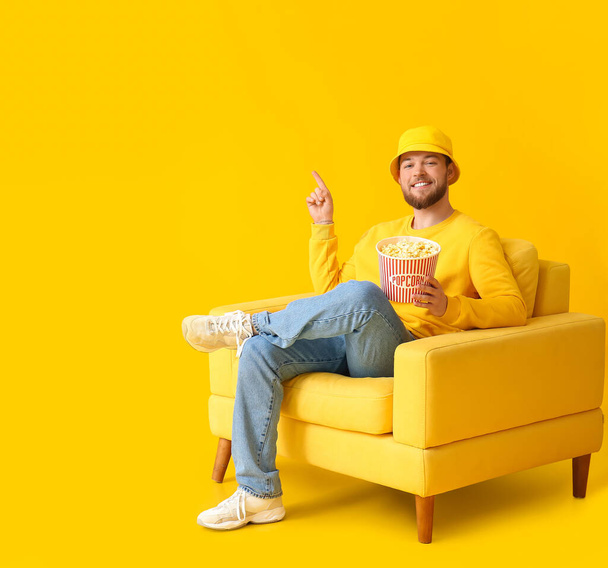 Junger Mann mit Popcorn sitzt im Sessel und zeigt auf etwas auf farbigem Hintergrund - Foto, Bild