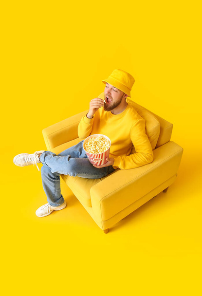Jongeman zittend in fauteuil en het eten van popcorn op kleur achtergrond - Foto, afbeelding