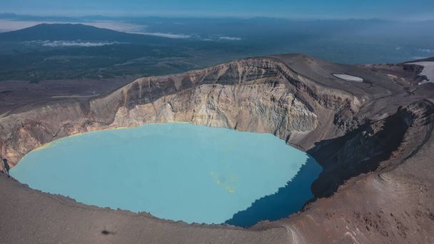 火山の火口にはターコイズブルーの生命のない酸性の湖がある。硫酸塩鉱床は水の上に見える。岩の多い海岸。斜面にヘリコプターの影。空中展望。カムチャツカ - 写真・画像