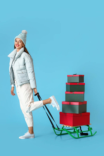 Beautiful young woman with sledge and Christmas gifts on blue background - 写真・画像