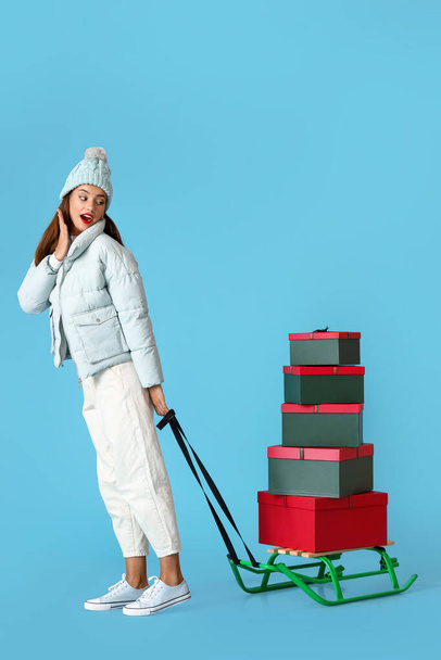 Surprised young woman with sledge and Christmas gifts on blue background - Фото, изображение