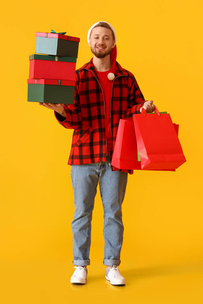 Fiatal férfi a Mikulás kalap karácsonyi ajándékok és táskák sárga háttér - Fotó, kép