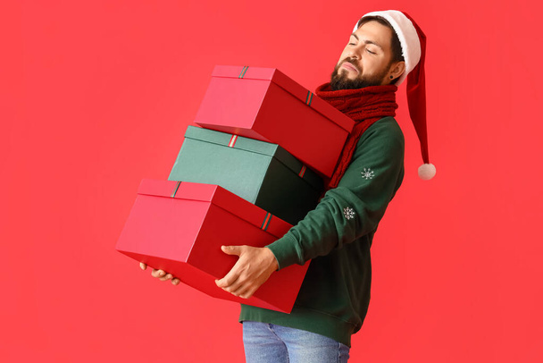 Handsome man in Santa hat with Christmas gifts on red background - Фото, изображение