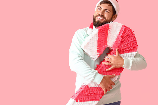 Hombre guapo en sombrero de Santa con piñata de bastón de caramelo sobre fondo rosa - Foto, Imagen