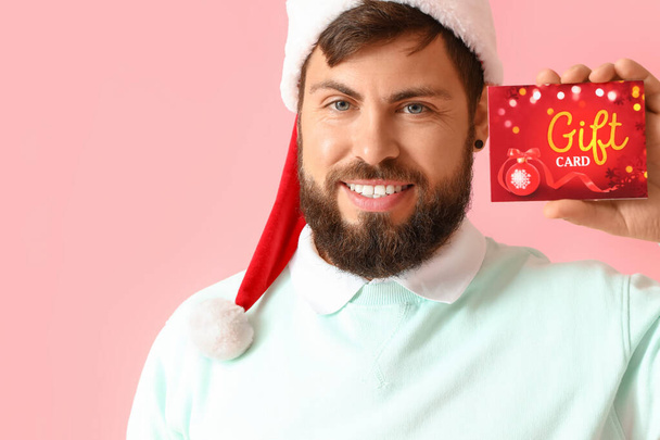 Handsome man in Santa hat with gift card on pink background - Fotó, kép