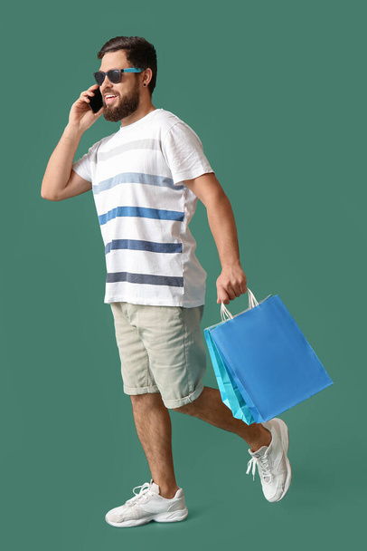 Boldog fiatal férfi fekete péntek bevásárló táskák beszél mobiltelefon zöld háttér - Fotó, kép