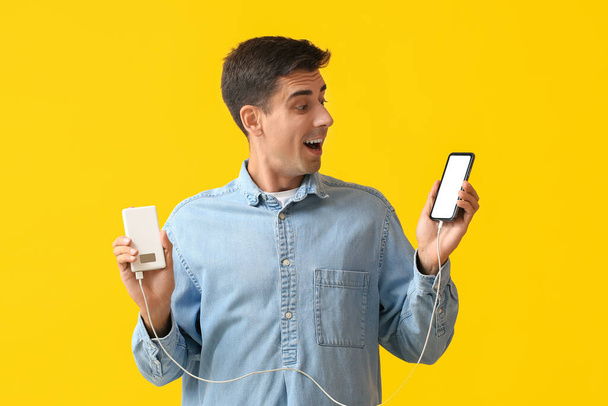Překvapený mladý muž s telefonem a power bank na barevném pozadí - Fotografie, Obrázek