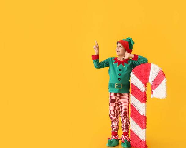 Lindo niño vestido como elfo con piñata de bastón de caramelo apuntando a algo en el fondo de color - Foto, Imagen