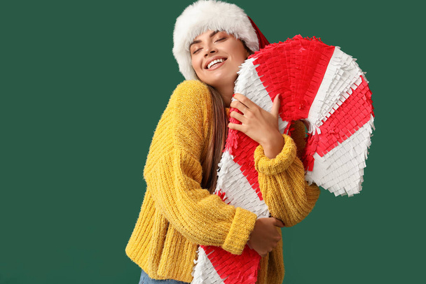 Pretty young woman in Santa hat with candy cane pinata on green background - Foto, Imagem