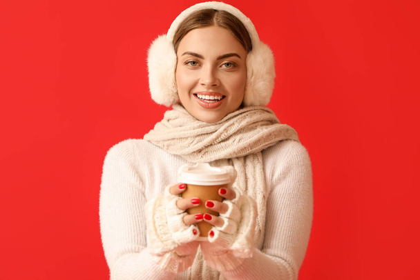 Hermosa mujer joven en orejeras calientes con una taza de café para llevar sobre fondo rojo - Foto, imagen