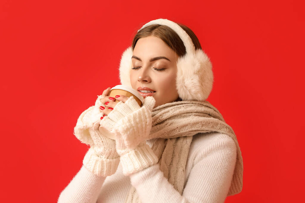 Beautiful young woman in warm earmuffs drinking coffee on red background - Фото, зображення