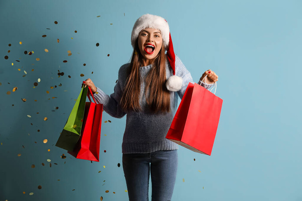 szczęśliwa młoda kobieta w kapeluszu Santa z torbami na zakupy i konfetti na niebieskim tle - Zdjęcie, obraz