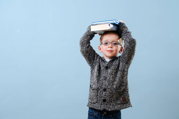 Playful little ginger boy in glasses holding books atop of his head. Over blue background. Studio shot. - 写真・画像