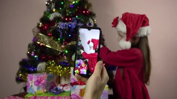 Noel Baba gibi giyinmiş küçük bir kız Noel ağacını süslüyor ve akıllı telefondan çekim yapmak için poz veriyor.. - Video, Çekim