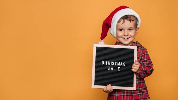Φωτογραφία στούντιο ενός αγοριού με χριστουγεννιάτικες πιτζάμες και ένα καπέλο σε κίτρινο φόντο με ένα πίνακα επιστολών με το κείμενο Καλά Χριστούγεννα στα χέρια του. Ένα μέρος για το κείμενό σας, διαφήμιση. - Φωτογραφία, εικόνα