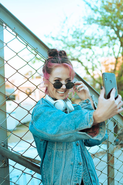 A vertical shot of a cool Caucasian lady with pink hair taking a selfie near a chain-link fence - Foto, imagen