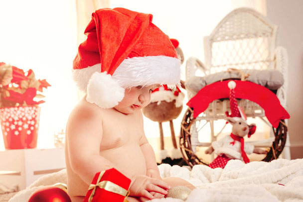 Beautiful baby child with santa claus hat and Christmas ornaments on the background. Happy little kid on a cozy blanket with balls and gift box. Merry Xmas and New Year holiday season concept - Φωτογραφία, εικόνα