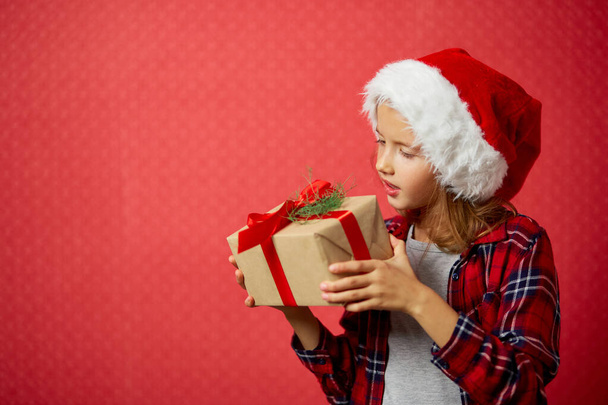 Zaskoczona i pozytywna dziewczynka w kapeluszu Mikołaja trzymając prezent świąteczny i sprawdzając pudełko prezentów, odizolowany na czerwonym tle, Szczęśliwego Nowego Roku - Zdjęcie, obraz