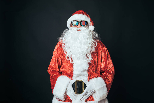 Homem vestido de Papai Noel usando óculos com carta de configuração de TV, em fundo preto. Conceito de Natal, Papai Noel, presentes, celebração. - Foto, Imagem