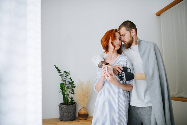 Weary thirty year old couple touching foreheads, holding coffee mugs. Man hugging woman from behind. He has a prosthetic bionic hand. - Photo, image