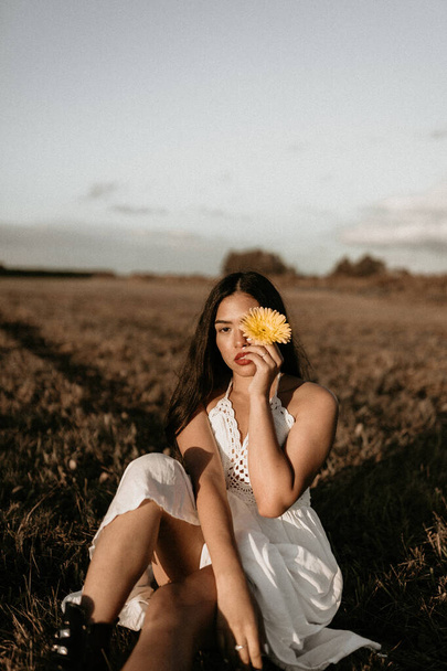 A cute brunette Hispanic woman posing in the field with a flower wearing a white elegant summer dress - 写真・画像