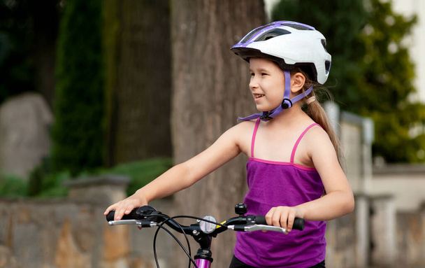 Mutlu ilkokul çocuğu kaskla bisiklete biniyor, bisikletli mutlu kız gülümsüyor, elleri demirli portrede, yakın plan. Bisiklet süren sportif bir çocuk, sıradan bir bisiklet. Genç bisikletçi portresi - Fotoğraf, Görsel