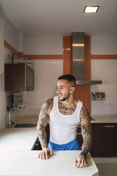 A vertical shot of a tattooed Caucasian male athlete standing at the kitchen - Foto, Imagem