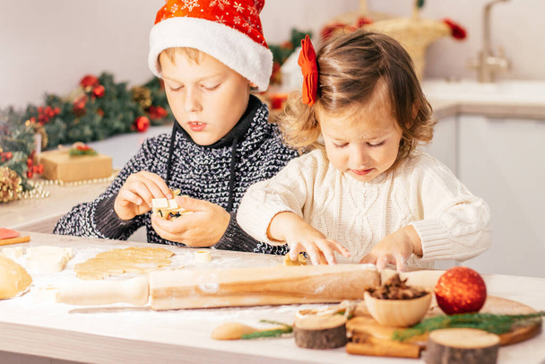 Χαριτωμένο κοριτσάκι 2-4 με κόκκινο φιόγκο και αγόρι 7-10 σε ένα χριστουγεννιάτικο καπέλο, κάνοντας χριστουγεννιάτικα μπισκότα μελόψωμο στην κουζίνα της Πρωτοχρονιάς. - Φωτογραφία, εικόνα