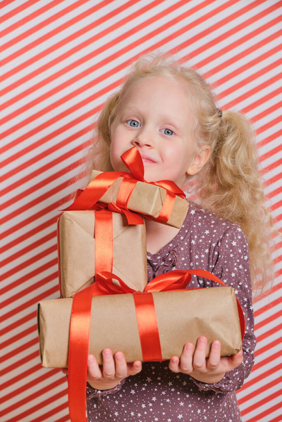 cute cheerful preschooler girl with gifts on a red striped background, gifts with a red ribbon in the hands of a girl with blond hair, boxing day, gifts for birthday and christmas, holiday gifts - Foto, Bild