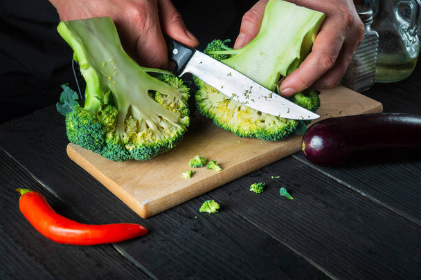 Cook cuts fresh Broccoli with a knife for salad on vintage kitchen table with fresh vegetables. Cooking and restaurant or cafe concept. - Photo, Image
