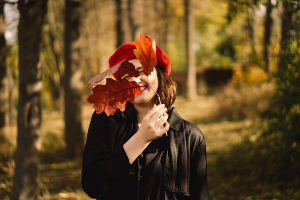 Teengirl v červeném baretu s kyticí podzimních listů v rukou kráčí lesem - Fotografie, Obrázek