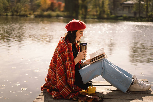 Uma adolescente em uma boina vermelha lendo livro sobre pontão de madeira. Temporada de outono. - Foto, Imagem